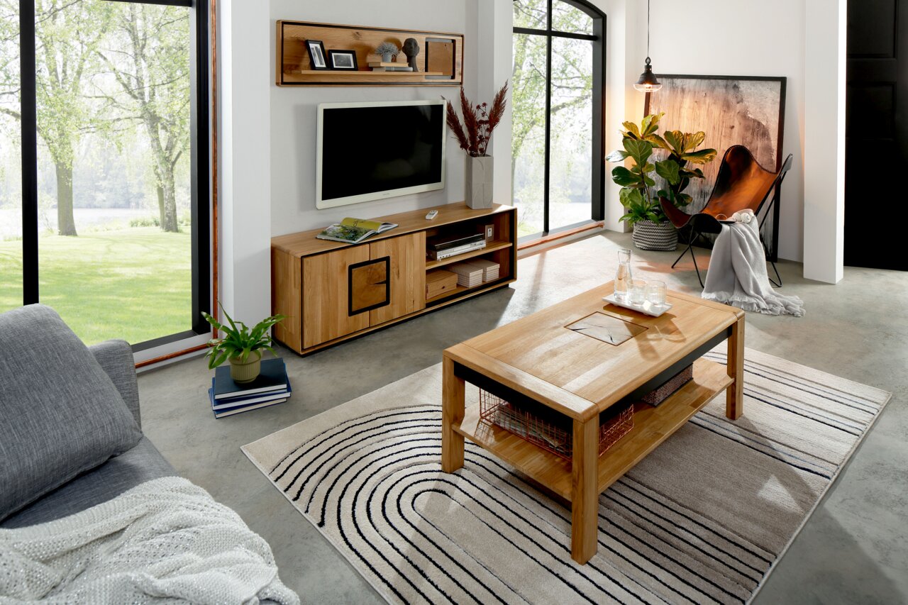 Modernes Wohnzimmer mit rechteckigem Couchtisch aus hellem Holz mit Ablagefach auf gemustertem Teppich, im Hintergrund TV-Board und Wandregal; Perspektive: leicht erhöhte, schräge Frontansicht von links.