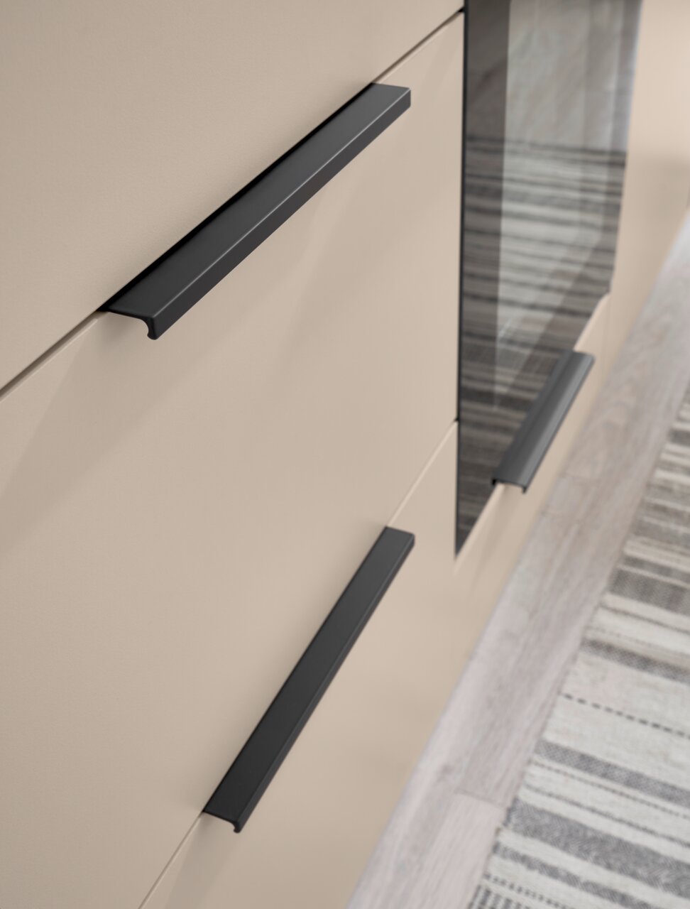 Detailaufnahme aus schräger Seitenperspektive: beige Schrankfront mit drei mattschwarzen, geradlinigen Griffleisten und einer dunklen Glas-/Spiegelfläche, daneben Teppich auf hellem Boden