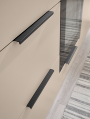 Detailaufnahme aus schräger Seitenperspektive: beige Schrankfront mit drei mattschwarzen, geradlinigen Griffleisten und einer dunklen Glas-/Spiegelfläche, daneben Teppich auf hellem Boden