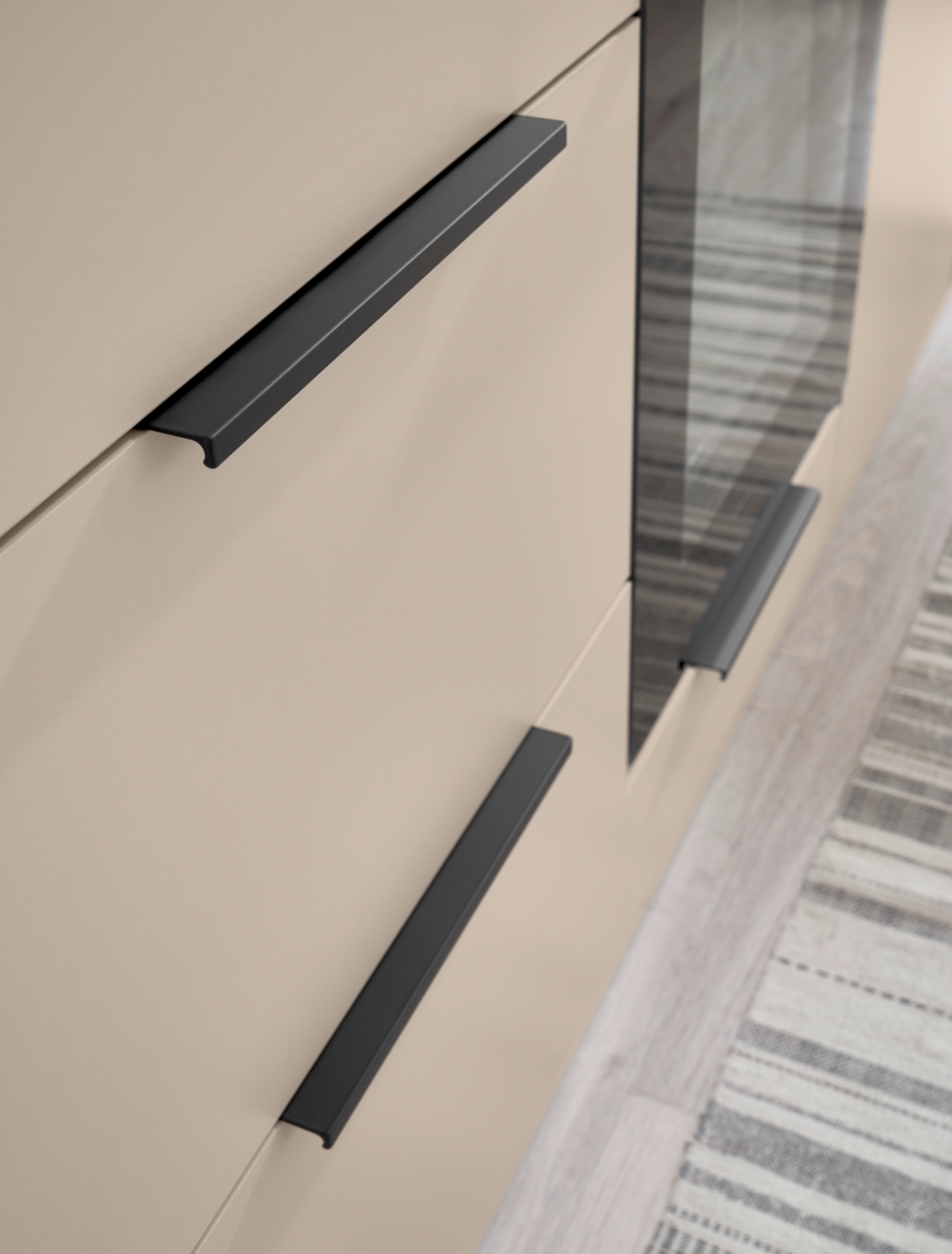 Detailaufnahme aus schräger Seitenperspektive: beige Schrankfront mit drei mattschwarzen, geradlinigen Griffleisten und einer dunklen Glas-/Spiegelfläche, daneben Teppich auf hellem Boden