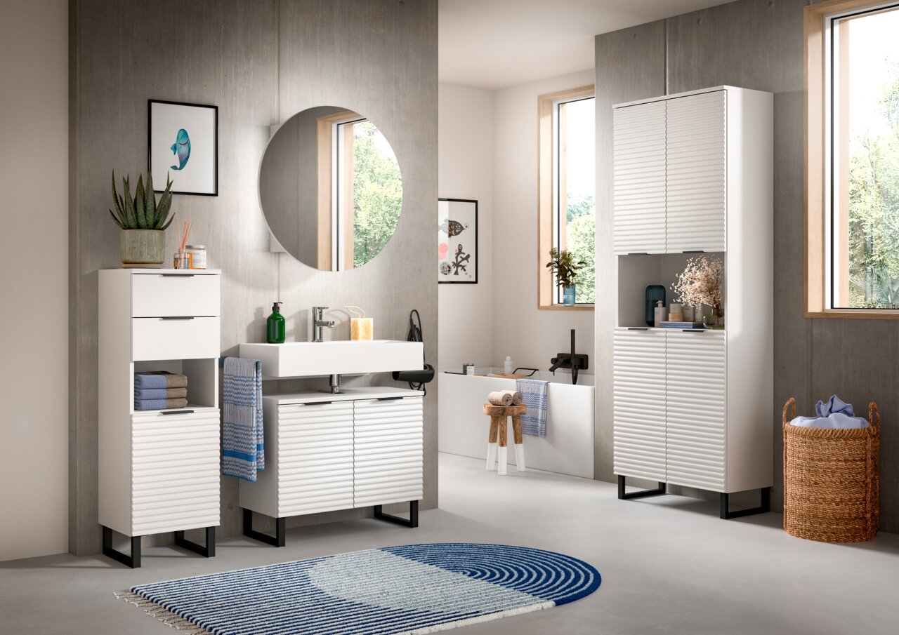 Moderner weißer Hochschrank im Badezimmer, frontal betrachtet, mit geriffelter Oberfläche und schwarzem Sockel, umgeben von weiteren Badmöbeln und Dekorationen.