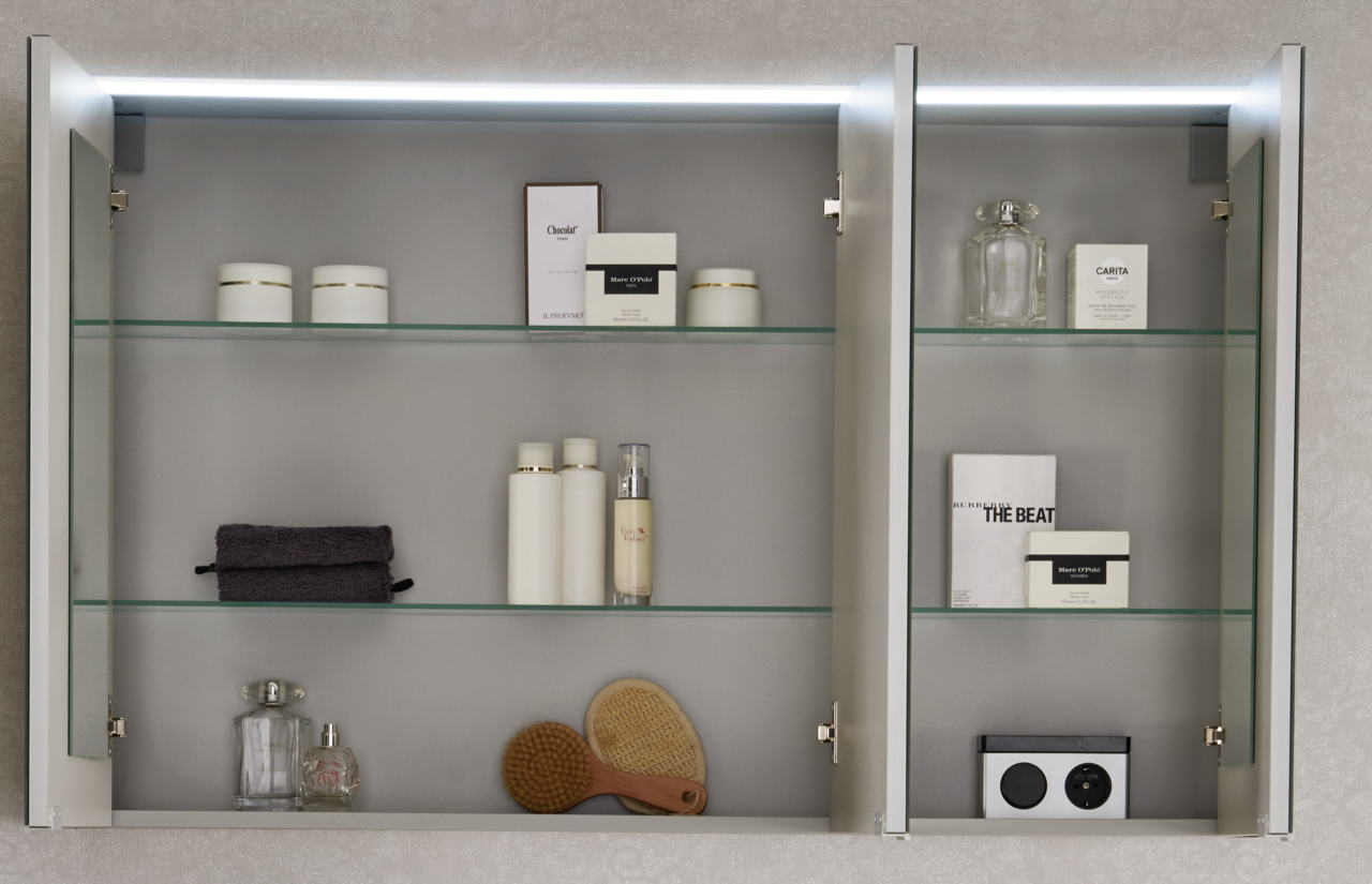 Geöffneter Spiegelschrank mit zwei Türen, beleuchtet von oben, gefüllt mit Pflegeprodukten und Accessoires, aus frontaler Perspektive.