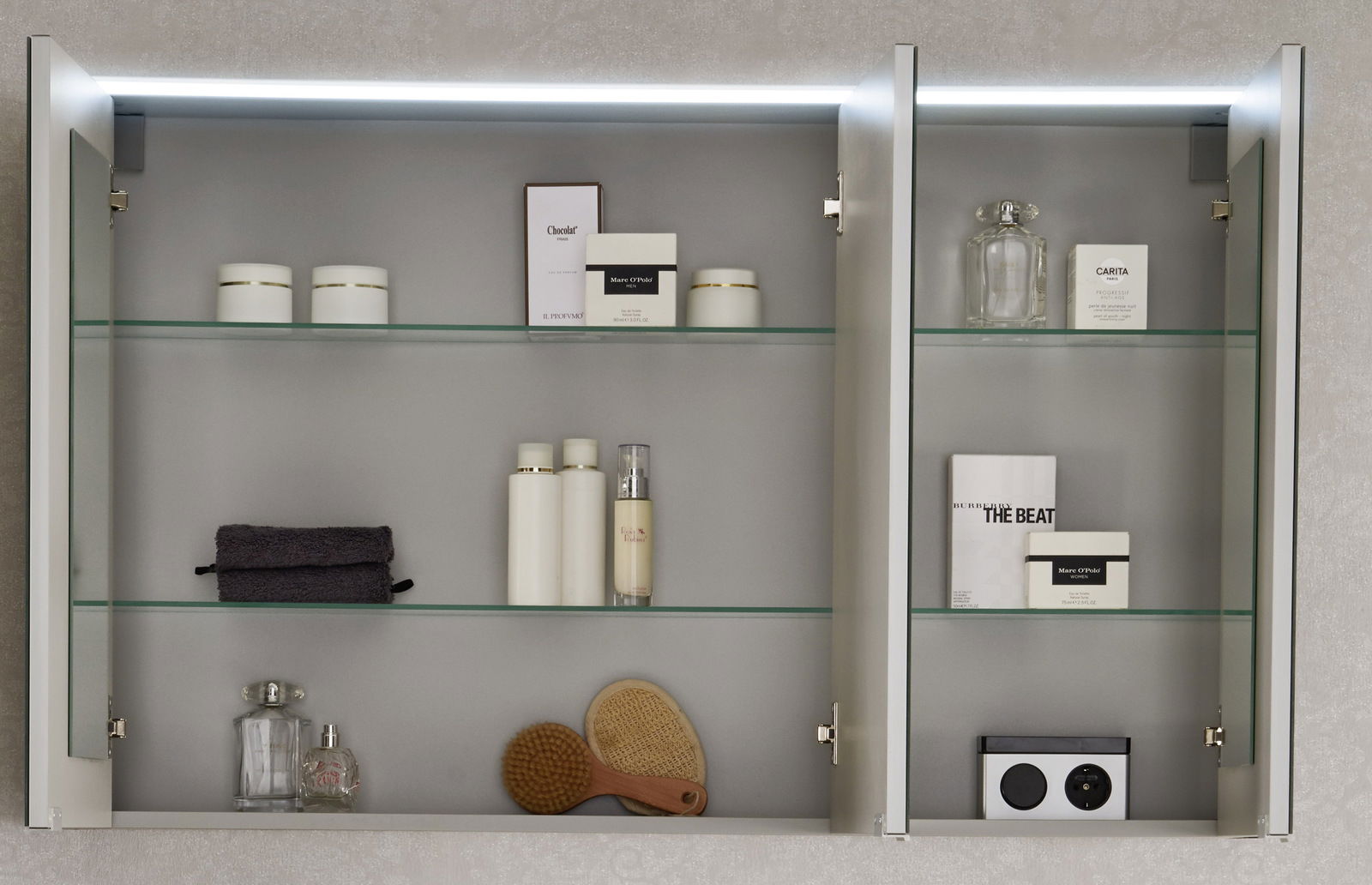 Geöffneter Spiegelschrank mit zwei Türen, beleuchtet von oben, gefüllt mit Pflegeprodukten und Accessoires, aus frontaler Perspektive.