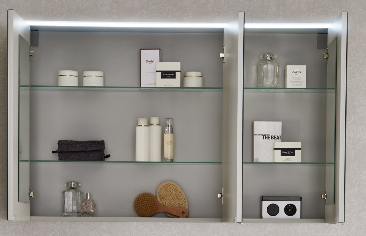 Geöffneter Spiegelschrank mit zwei Türen, beleuchtet von oben, gefüllt mit Pflegeprodukten und Accessoires, aus frontaler Perspektive.