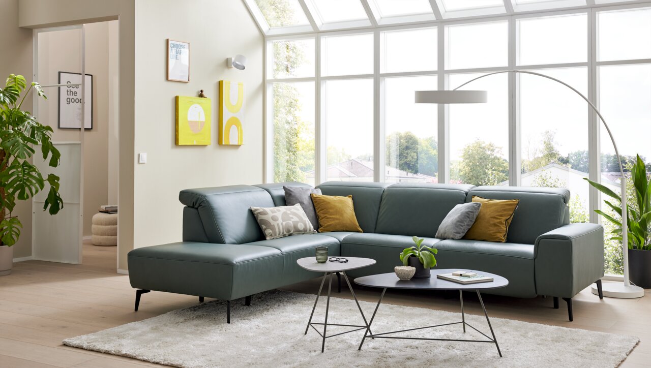 Moderne hoekbank in een lichte woonkamer met grote ramen, groene planten en stijlvolle decoraties, gezien vanuit de zijkant.
