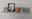 Graues Wandregal mit Dekoration, darunter ein gerahmtes Bild, ein orangefarbener Übertopf und eine Kerze in einem Glas. Perspektive von vorne.