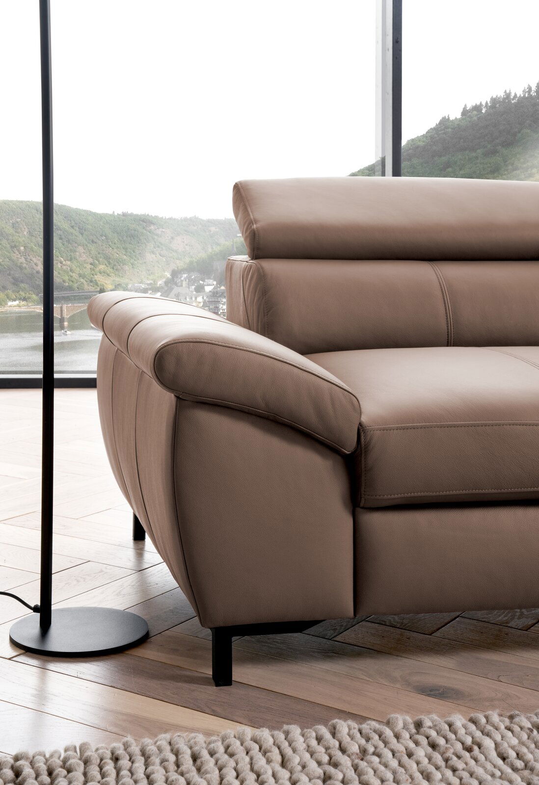 Nahaufnahme einer braunen Ledercouch mit Armlehne und verstellbarer Kopfstütze, im Hintergrund ein großes Fenster mit Blick auf eine grüne Landschaft.