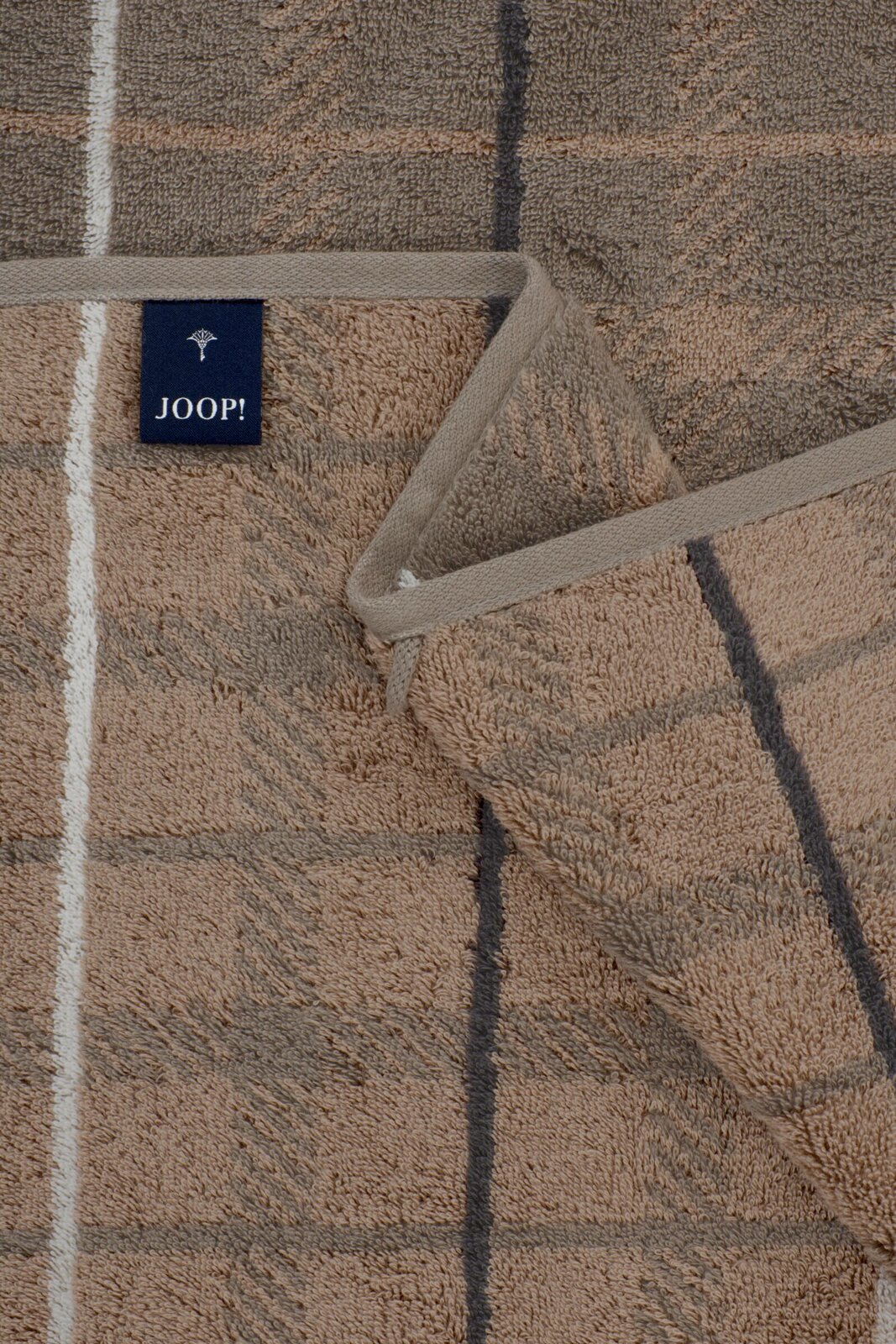 Nahaufnahme aus leicht schräger Draufsicht auf ein JOOP! Frottee-Handtuch in Beige/Taupe mit geometrischem Linienmuster und umlaufender Einfassung