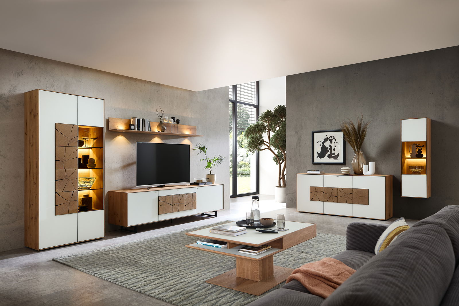 Moderne Wohnzimmer-Wandkombination in Holzoptik mit weißen Fronten, bestehend aus einem TV-Schrank, einem hohen Schrank und einem Hängeschrank, mit dekorativer Beleuchtung und stilvollen Accessoires, seitliche Perspektive.