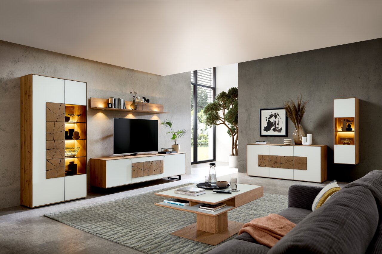 Valnatura Wandcombinatie LAGO Moderne Wohnzimmer-Wandkombination in Holzoptik mit weißen Fronten, bestehend aus einem TV-Schrank, einem hohen Schrank und einem Hängeschrank, mit dekorativer Beleuchtung und stilvollen Accessoires, seitliche Perspektive.