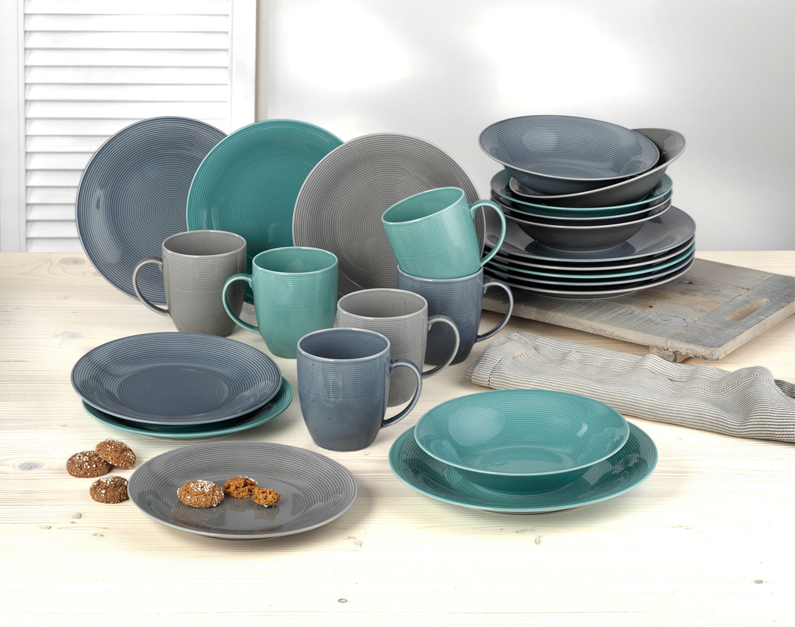 Kombiservice 24-teilig Jive in verschiedenen Grautönen und Türkis, bestehend aus Tellern, Schalen und Tassen, auf einem hellen Holztisch aus der Vorderansicht.