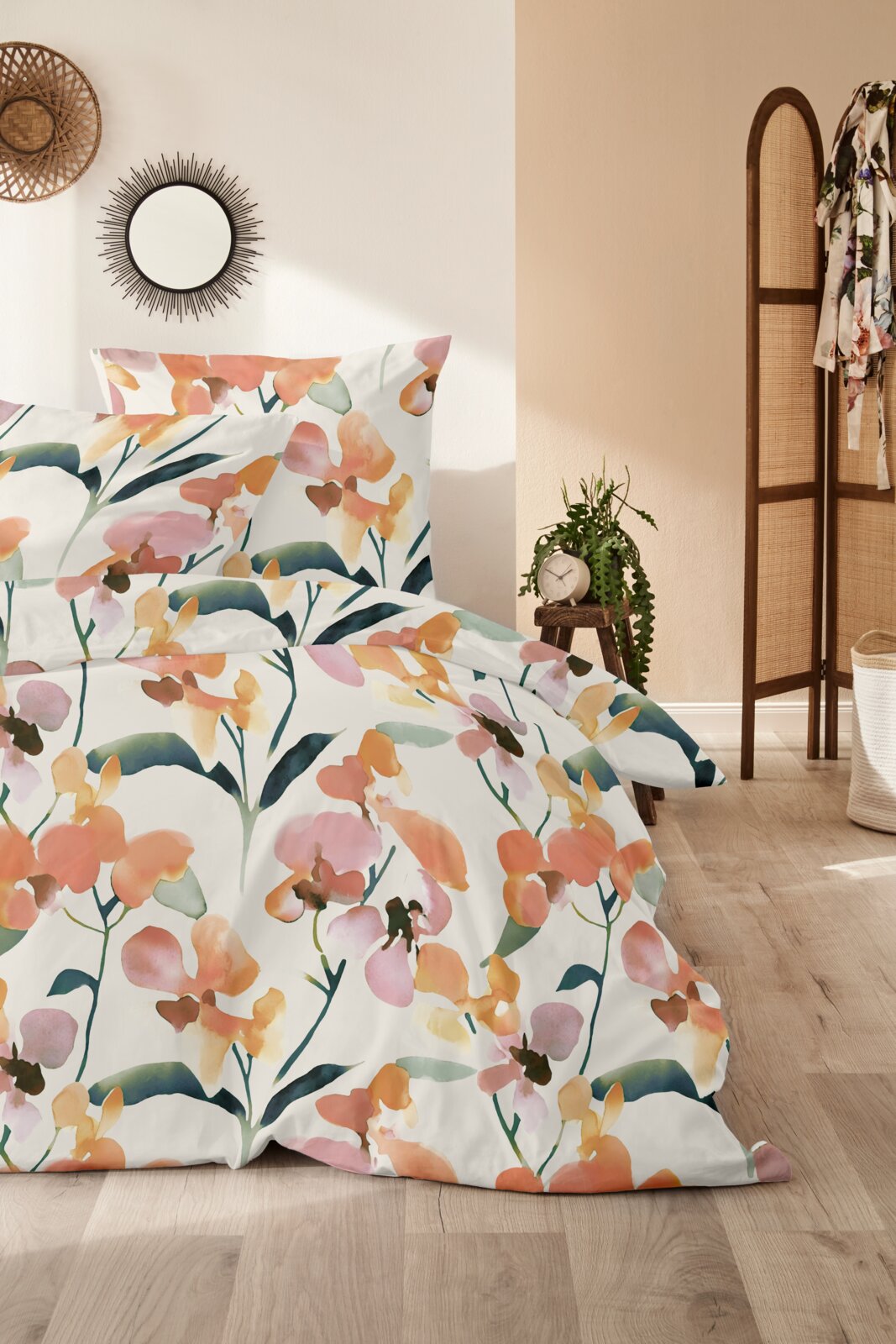 Blumige Bettwäsche mit Aquarell-Blütenmuster in Orange, Rosa und Grün auf weißem Grund, frontal leicht seitlich fotografiert im hellen Schlafzimmer-Setting.