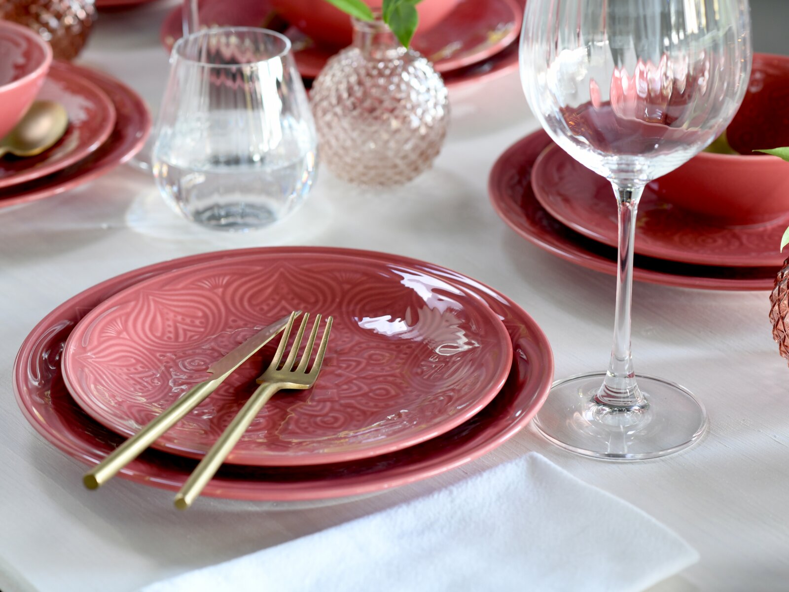Schräg von oben fotografiertes, festlich gedecktes Tischarrangement mit roséfarbenen, geprägten Keramiktellern, goldfarbenem Besteck und Weingläsern auf weißer Tischdecke