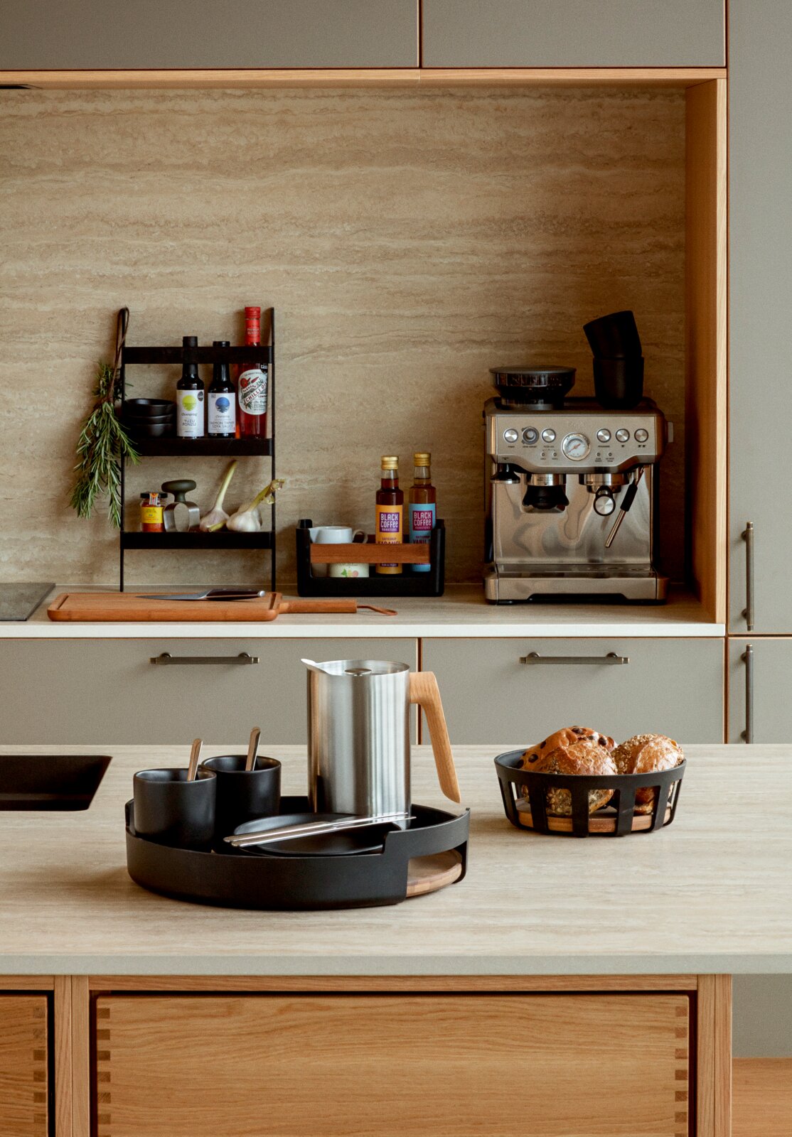 Stijlvolle keukenopstelling met zwarte serveertray met kopjes en zilverkleurige koffiekan op het aanrecht, naast een broodmand en een espressomachine op de achtergrond, gefotografeerd in frontaal perspectief op ooghoogte.