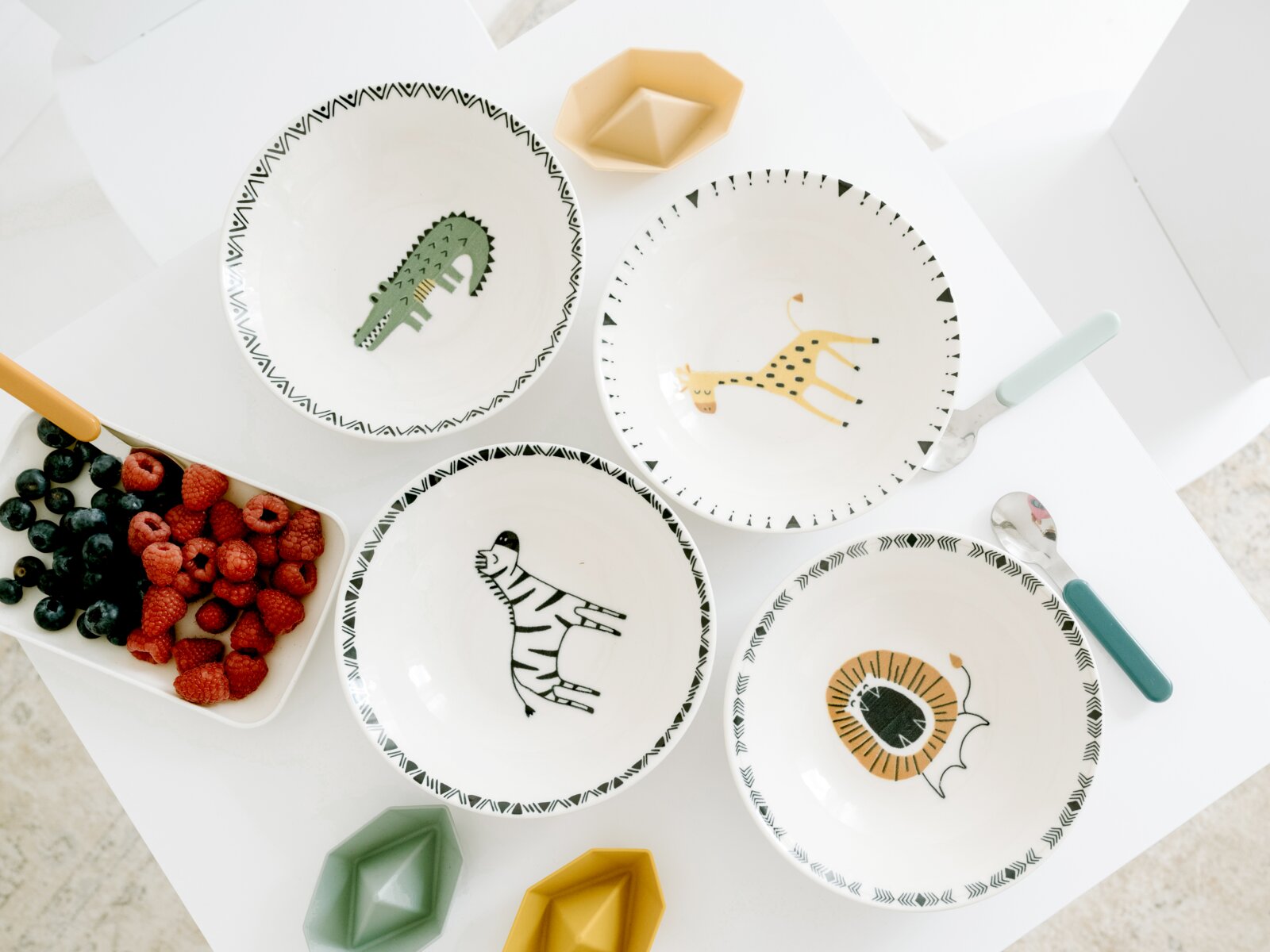 creaTable Schalen-Set 4-tlg. SAFARI Draufsicht auf vier weiße Kinderschalen mit schwarz gemustertem Rand und Tiermotiven (Krokodil, Giraffe, Zebra, Löwe) auf einem weißen Tisch, daneben Beeren und Löffel mit bunten Griffen