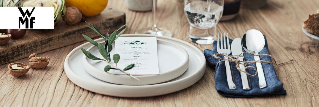 Ein festlich gedeckter Holztisch mit weißem Geschirr, Besteck auf Stoffserviette und dekorativen Nüssen.