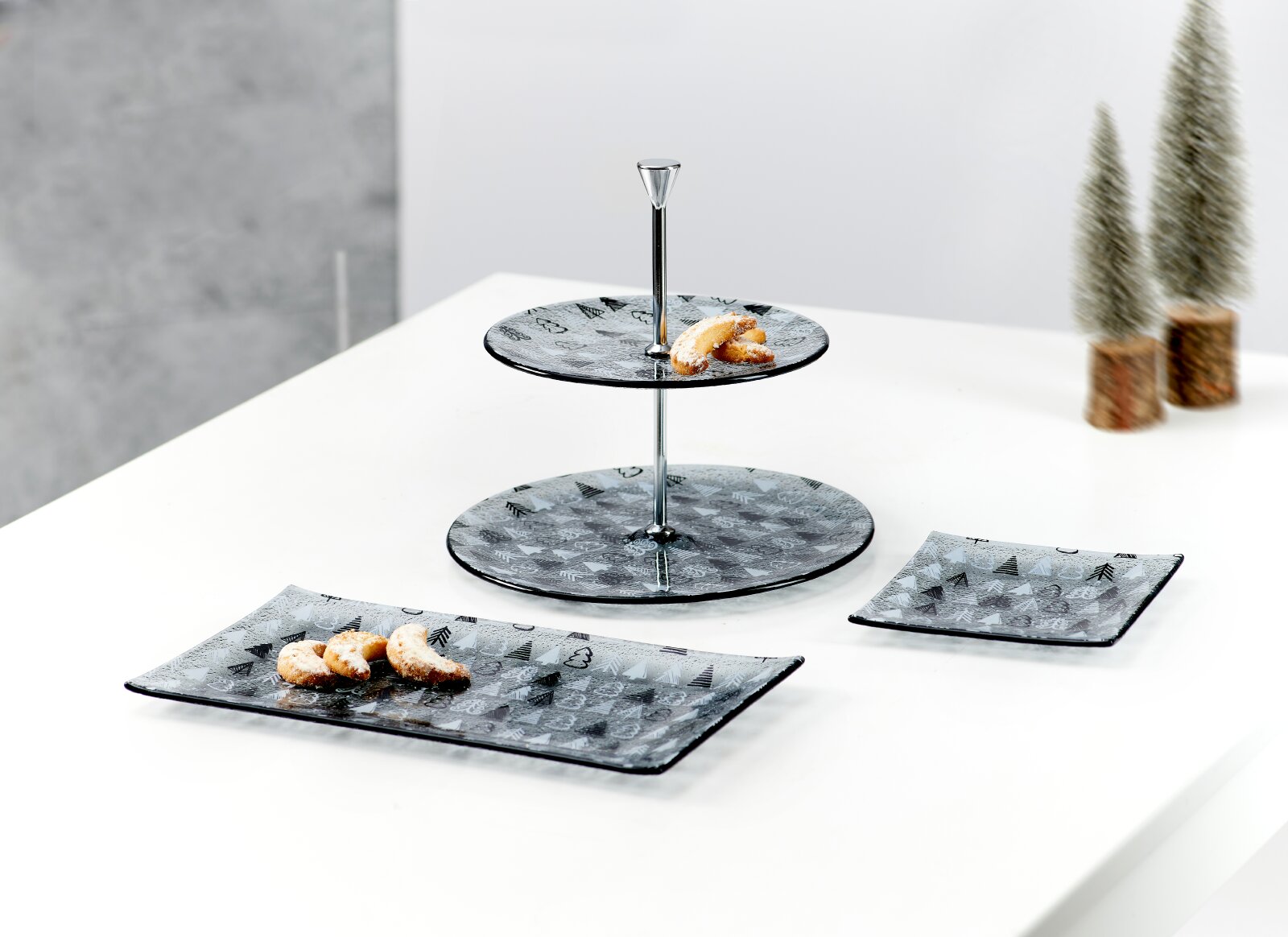 Zweistöckige Etagere und zwei Servierplatten aus grauem Glas mit winterlichem Muster, dekoriert mit Gebäck, aus schräger Vorderansicht