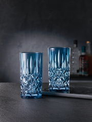 Zwei elegante Longdrinkgläser aus der Noblesse-Serie in blauer Kristalloptik, frontal auf einem dunklen Hintergrund präsentiert.