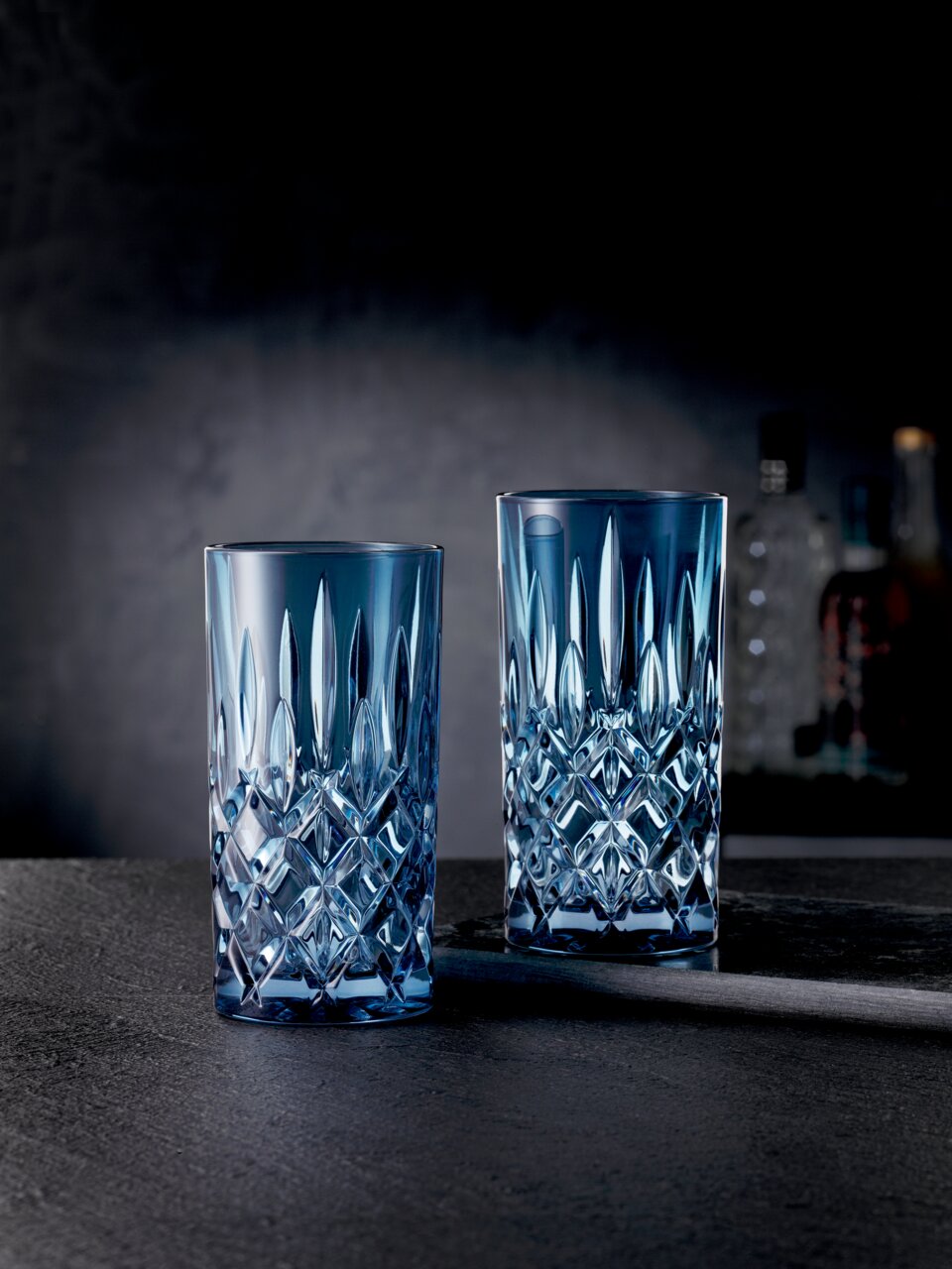 Zwei elegante Longdrinkgläser aus der Noblesse-Serie in blauer Kristalloptik, frontal auf einem dunklen Hintergrund präsentiert.