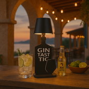 Stimmungsvolle Akku-Tischleuchte in Form einer Gin-Flasche mit der Aufschrift 'Gintastic', im Vordergrund ein Glas mit Gin Tonic und Limettenscheiben, im Hintergrund ein Sonnenuntergang auf einer Terrasse.
