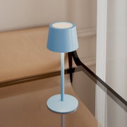 Hellblaue, minimalistische Tischlampe mit zylindrischem Schirm auf rundem Standfuß, auf einem Glastisch neben einem beigen Sessel, in leicht erhöhter Frontansicht.
