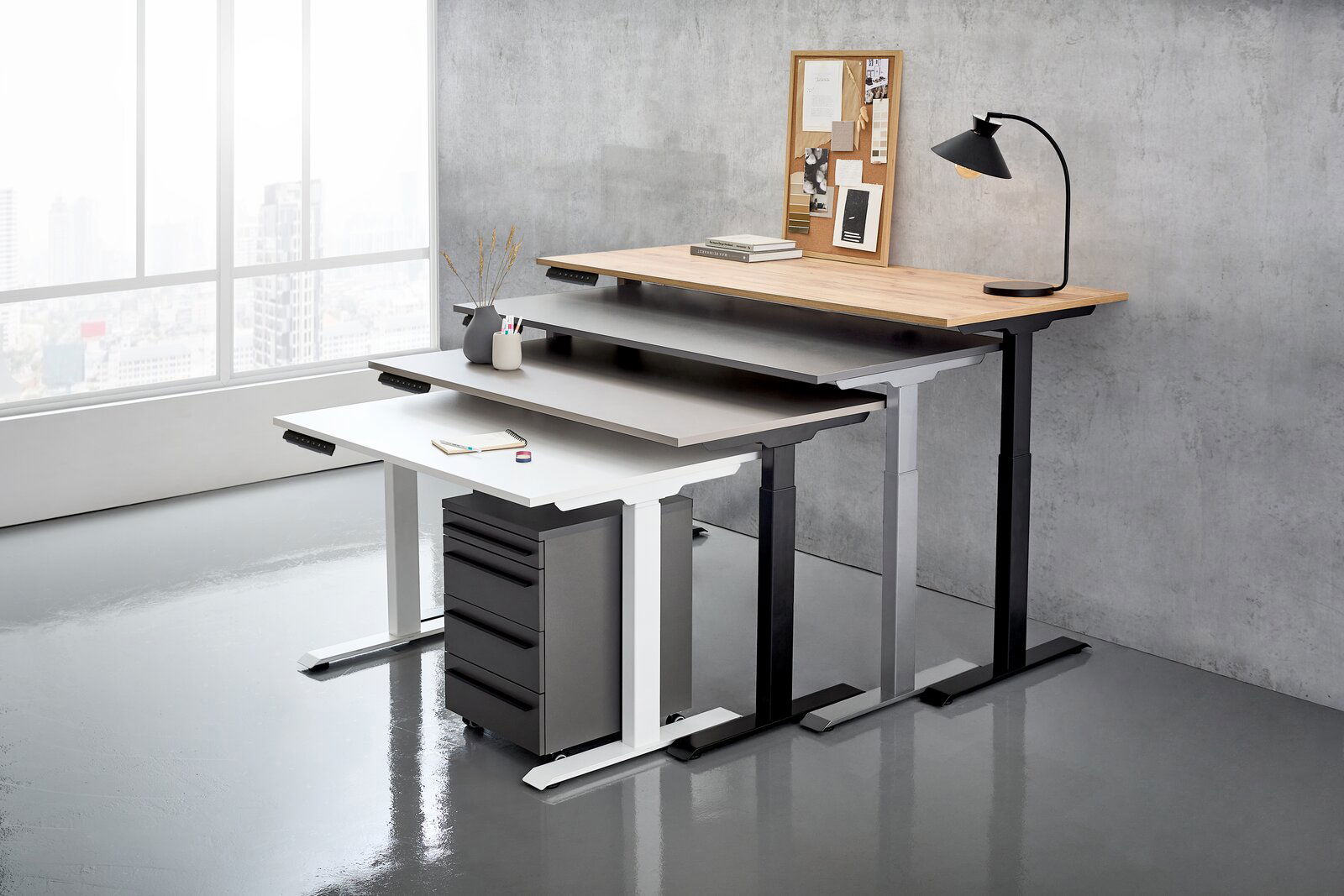 Drei höhenverstellbare E-Schreibtische in verschiedenen Farben und Größen, gestapelt in einem modernen Büro mit Blick auf die Stadt durch ein großes Fenster.