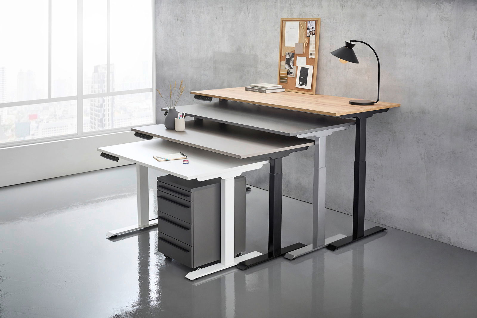 Drei höhenverstellbare E-Schreibtische in verschiedenen Farben und Größen, gestapelt in einem modernen Büro mit Blick auf die Stadt durch ein großes Fenster.