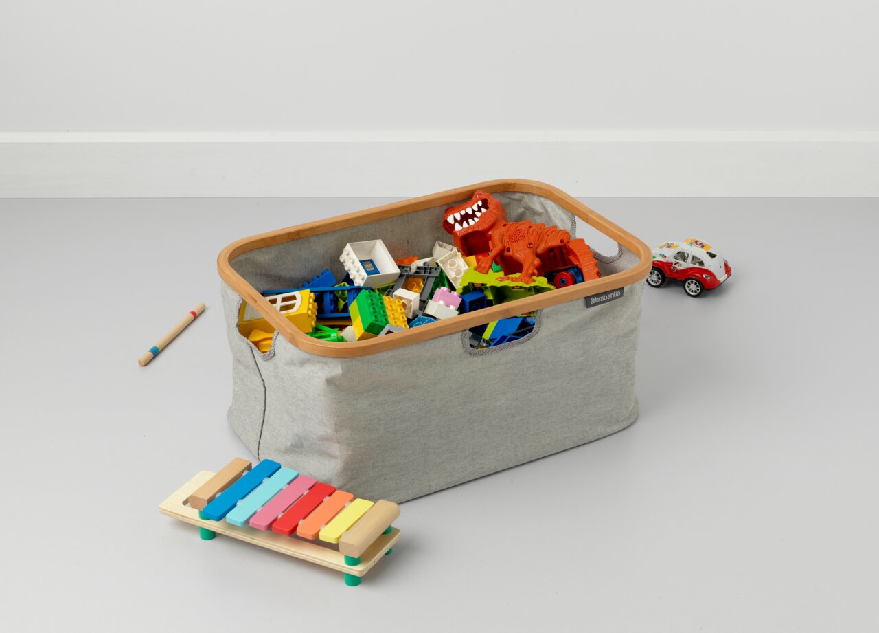 Opvouwbare wasmand van grijze stof met houten frame, gevuld met kleurrijk speelgoed, gefotografeerd vanuit een zijaanzicht.