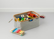Opvouwbare wasmand van grijze stof met houten frame, gevuld met kleurrijk speelgoed, gefotografeerd vanuit een zijaanzicht.