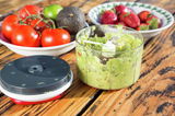 Easy Pull Universalhacker mit abgenommenem Deckel, gefüllt mit frisch zubereitetem Guacamole, auf einem Holztisch. Im Hintergrund Schalen mit Tomaten, Avocado, Limette und Erdbeeren.