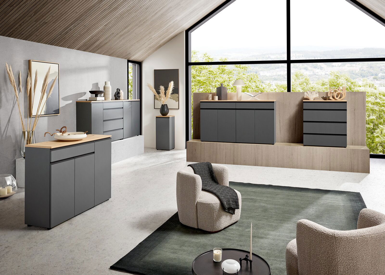 Dieter Knoll XL-Sideboard LENDINA Moderne Wohnzimmermöbel in einem hellen Raum mit großen Fenstern, die eine grüne Aussicht bieten. Die Möbel sind in einem eleganten, dunklen Grau gehalten und umfassen Sideboards und Kommoden. Die Perspektive zeigt den Raum von schräg vorne, mit einem gem