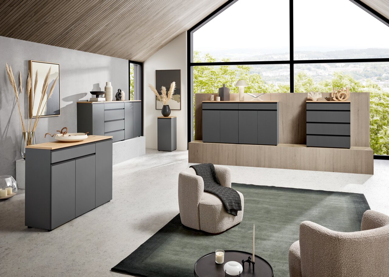 Moderne Wohnzimmermöbel in einem hellen Raum mit großen Fenstern, die eine grüne Aussicht bieten. Die Möbel sind in einem eleganten, dunklen Grau gehalten und umfassen Sideboards und Kommoden. Die Perspektive zeigt den Raum von schräg vorne, mit einem gem
