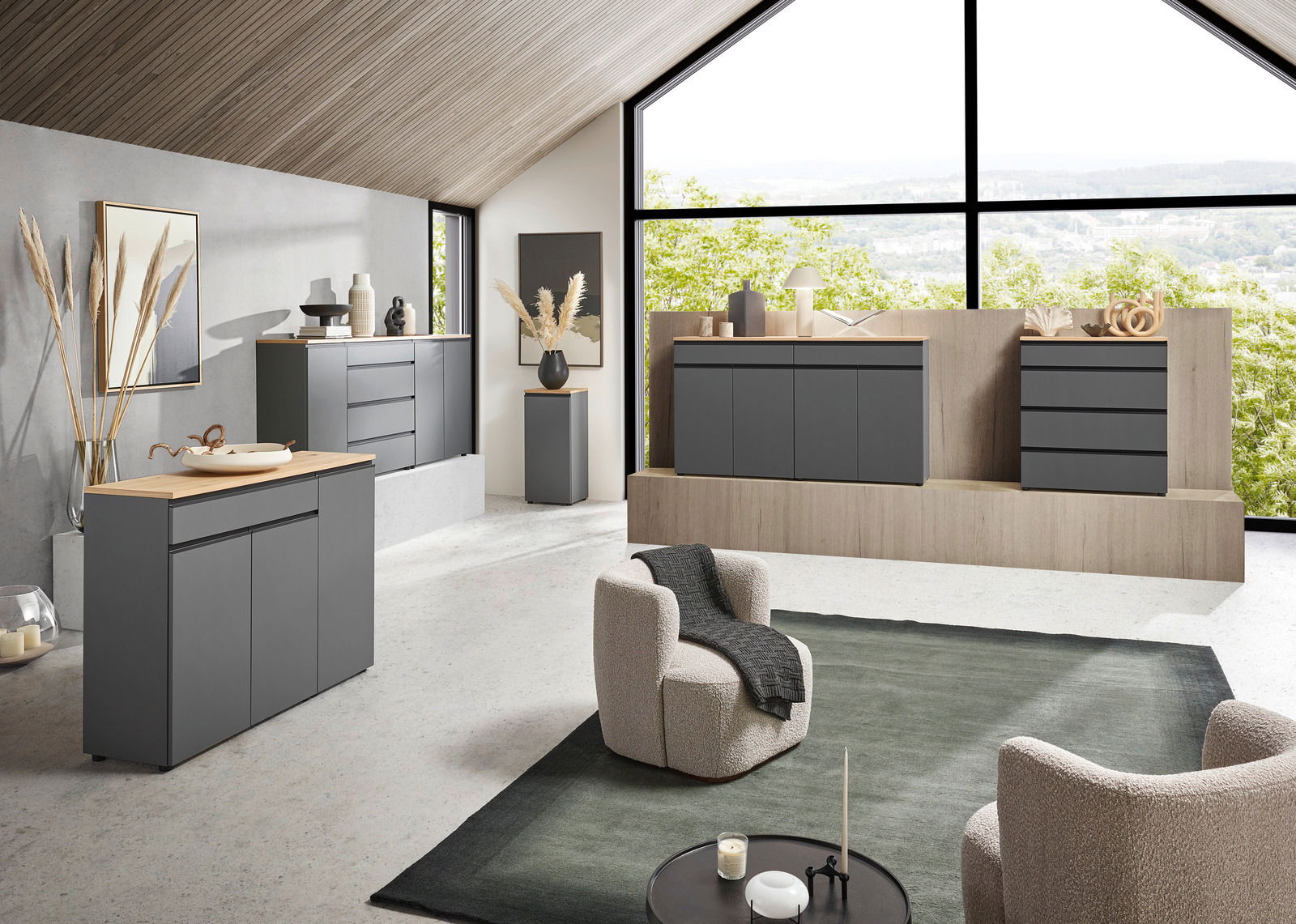Moderne Wohnzimmermöbel in einem hellen Raum mit großen Fenstern, die eine grüne Aussicht bieten. Die Möbel sind in einem eleganten, dunklen Grau gehalten und umfassen Sideboards und Kommoden. Die Perspektive zeigt den Raum von schräg vorne, mit einem gem