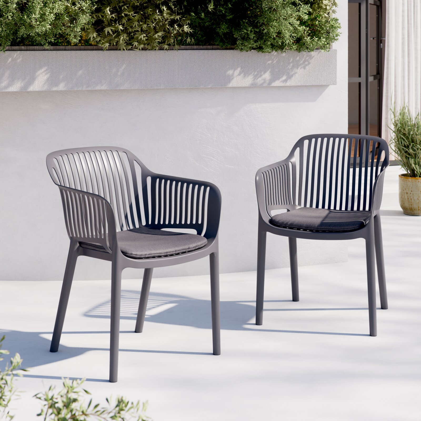 Zwei moderne graue Outdoor-Stühle aus Kunststoff mit Sitzkissen auf einer hellen Terrasse vor weißer Wand und Pflanzen, Perspektive: frontale Ansicht leicht von links