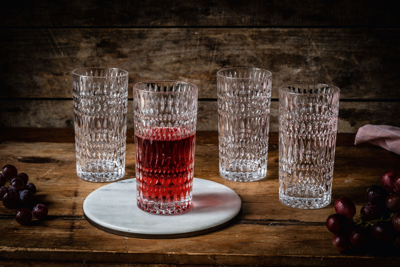 Vier Longdrinkgläser im Ethno-Stil auf einem Holztisch, eines gefüllt mit rotem Getränk, aus frontaler Perspektive.