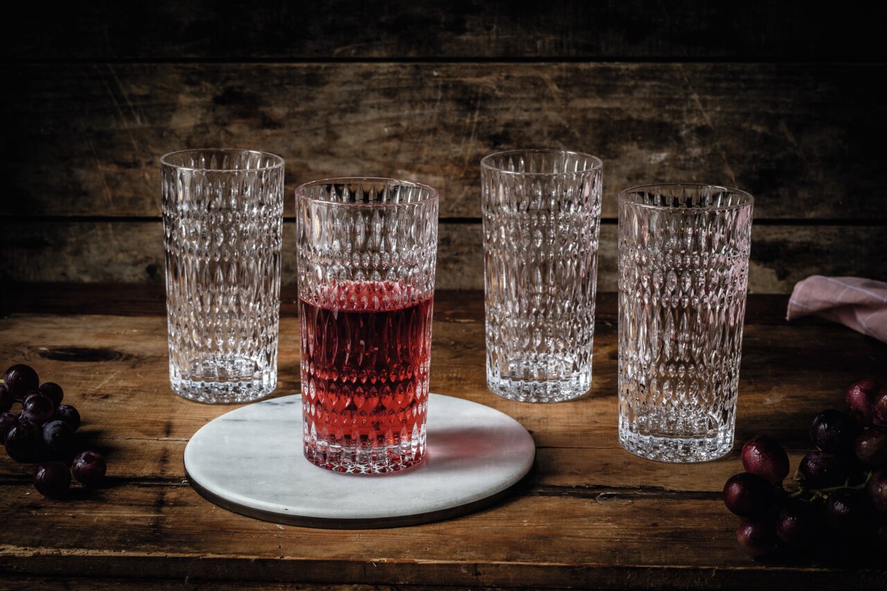 Vier Longdrinkgläser im Ethno-Stil auf einem Holztisch, eines gefüllt mit rotem Getränk, aus frontaler Perspektive.