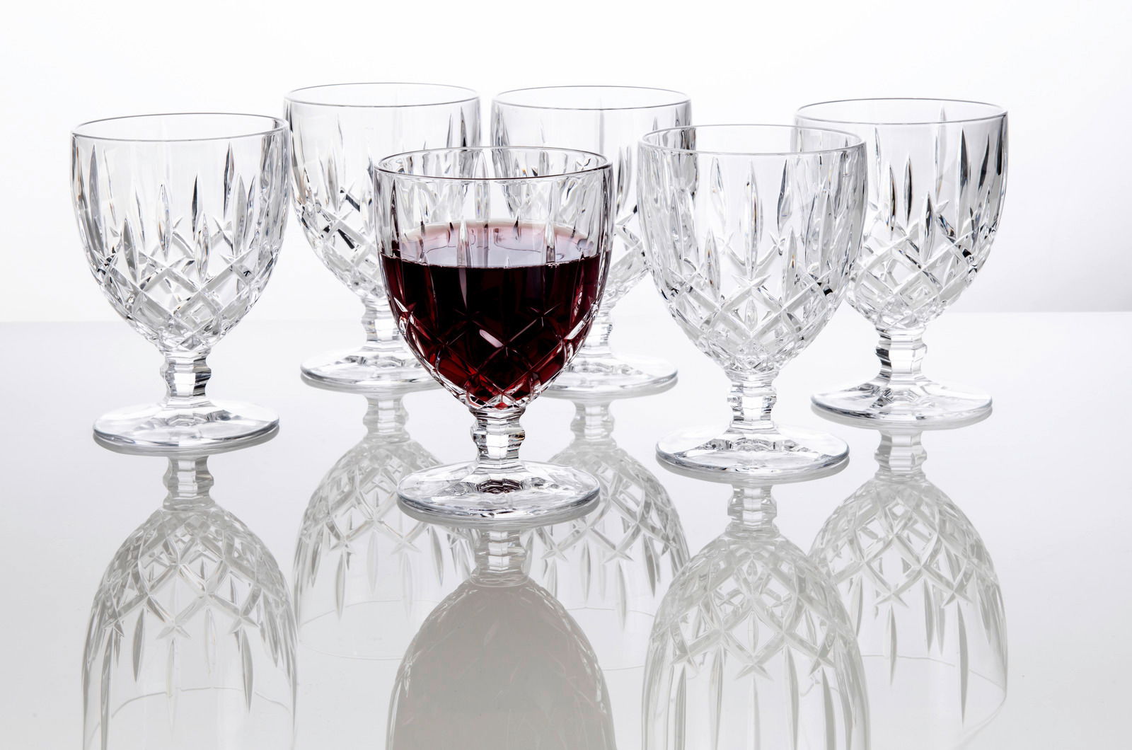 Sechs elegante Weinkelche Noblesse 350ml aus Kristallglas, einer davon mit Rotwein gefüllt, auf einer reflektierenden Oberfläche, seitliche Perspektive.