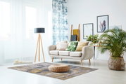 Moderner Wohnzimmerteppich in Grau- und Brauntönen, zentral im Raum platziert, mit einem runden, geflochtenen Pouf darauf. Perspektive von vorne.