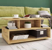 Moderner Couchtisch aus hellem Holz mit offenem Stauraum und Ablagefächern, dekoriert mit Tassen, Schale und Büchern vor grünem Sofa, perspektivisch in leichter Schrägansicht von vorn.