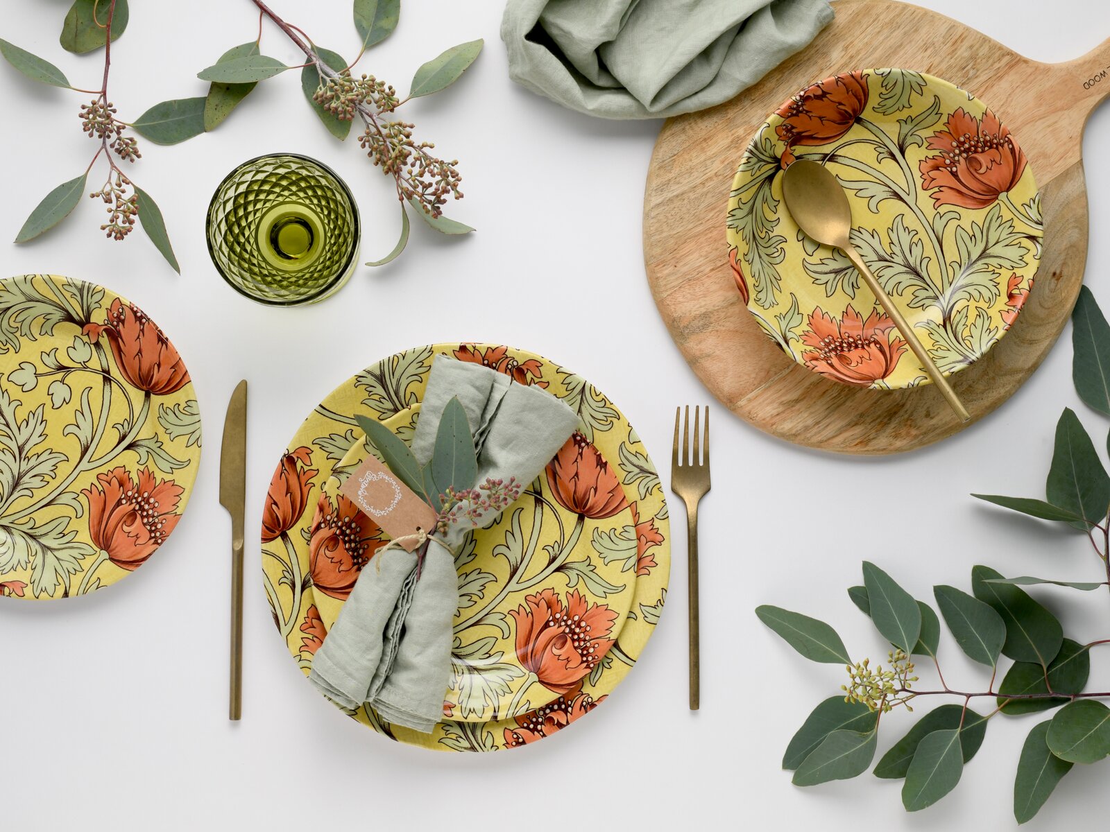 Draufsicht (Flatlay) auf stilvolles Tischgedeck mit gelben Tellern im floralen Muster in Orange und Grün, goldenem Besteck, grünem Trinkglas, Leinenserviette und Eukalyptuszweigen auf weißem Hintergrund