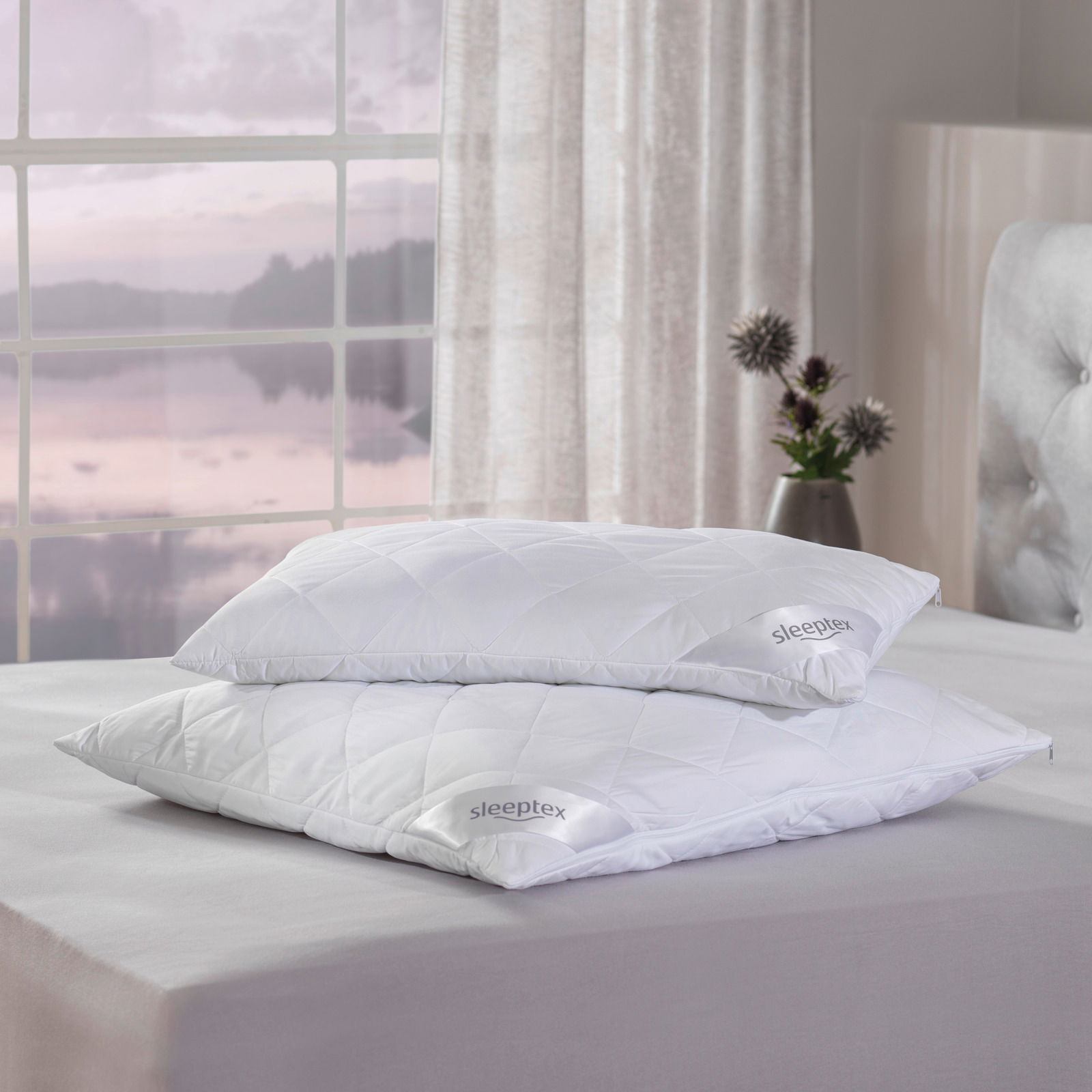 Zwei weiße Sleeptex Kopfkissen OVIEDO auf einem Bett, seitliche Perspektive mit einem Fenster im Hintergrund.