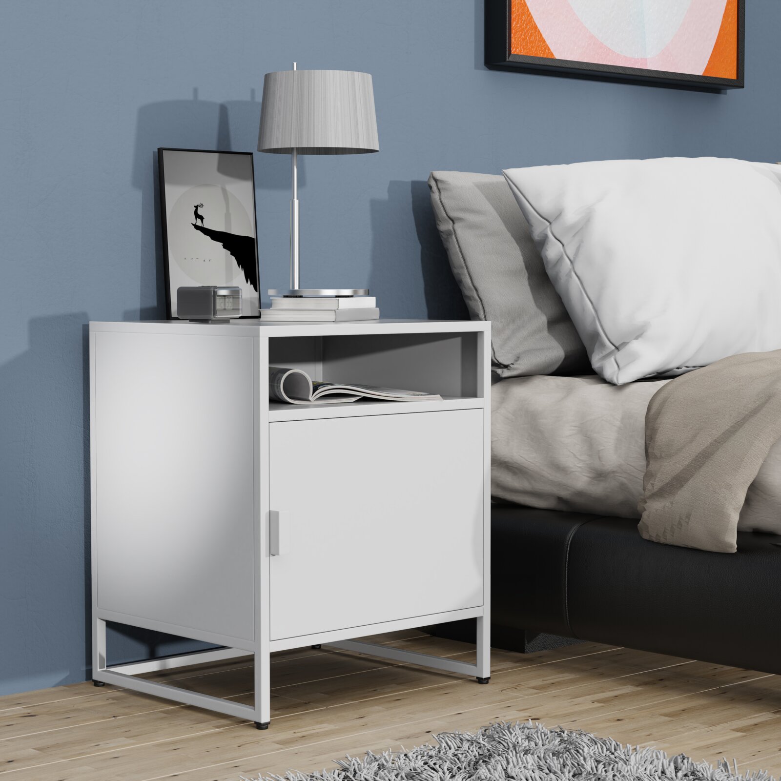 Weißer moderner Nachttisch mit Türfach und offenem Ablagefach neben einem Bett, darauf Lampe und Deko, aufgenommen aus leicht schräger Frontperspektive auf Augenhöhe.