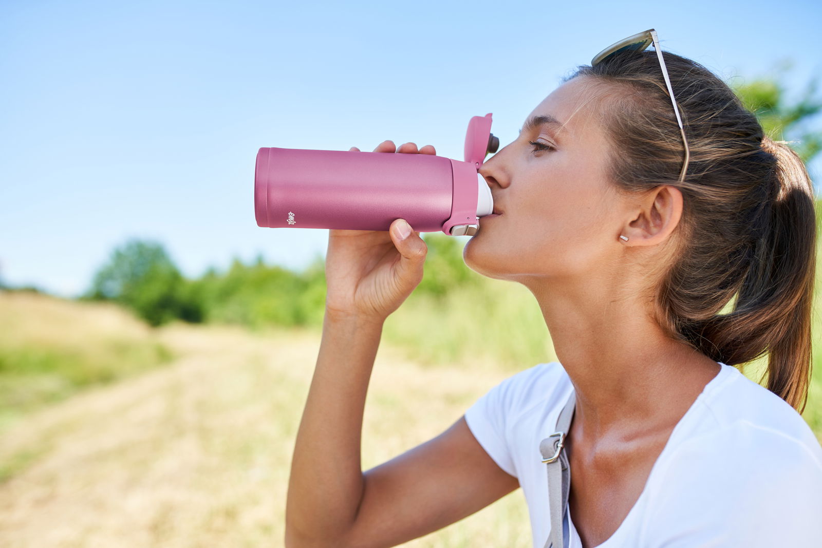 Frau trinkt aus einem rosa Thermobecher in der Natur, seitliche Perspektive