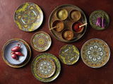 Vogelperspektive auf ein 12-teiliges Tellerset mit Mandala-Muster in lebendigen Farben, arrangiert auf einem Tisch mit Gewürzen und Gemüse.
