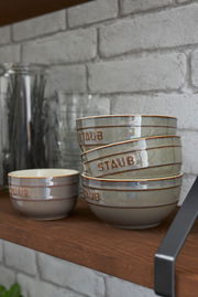 Graues Schüsselset, bestehend aus vier stapelbaren Schüsseln mit der Aufschrift 'STAUB', auf einem Holzregal vor einer Ziegelwand.