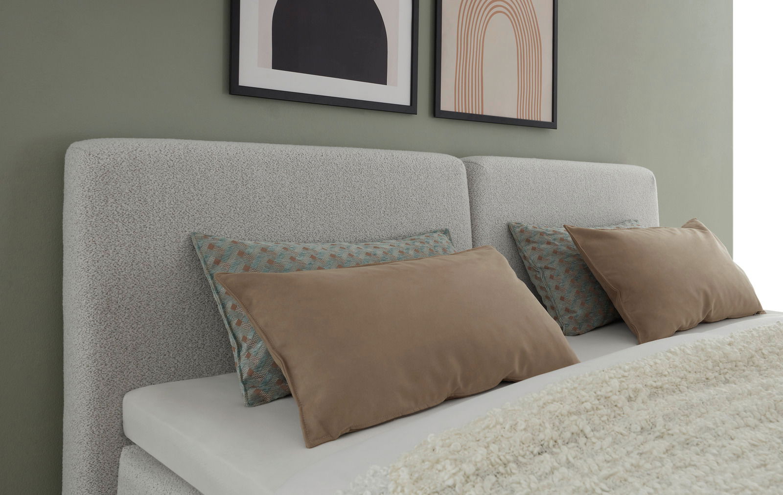 Nahaufnahme eines Boxspringbetts mit grauem Kopfteil, dekoriert mit Kissen in Erdtönen und gemusterten Bezügen, seitliche Perspektive.