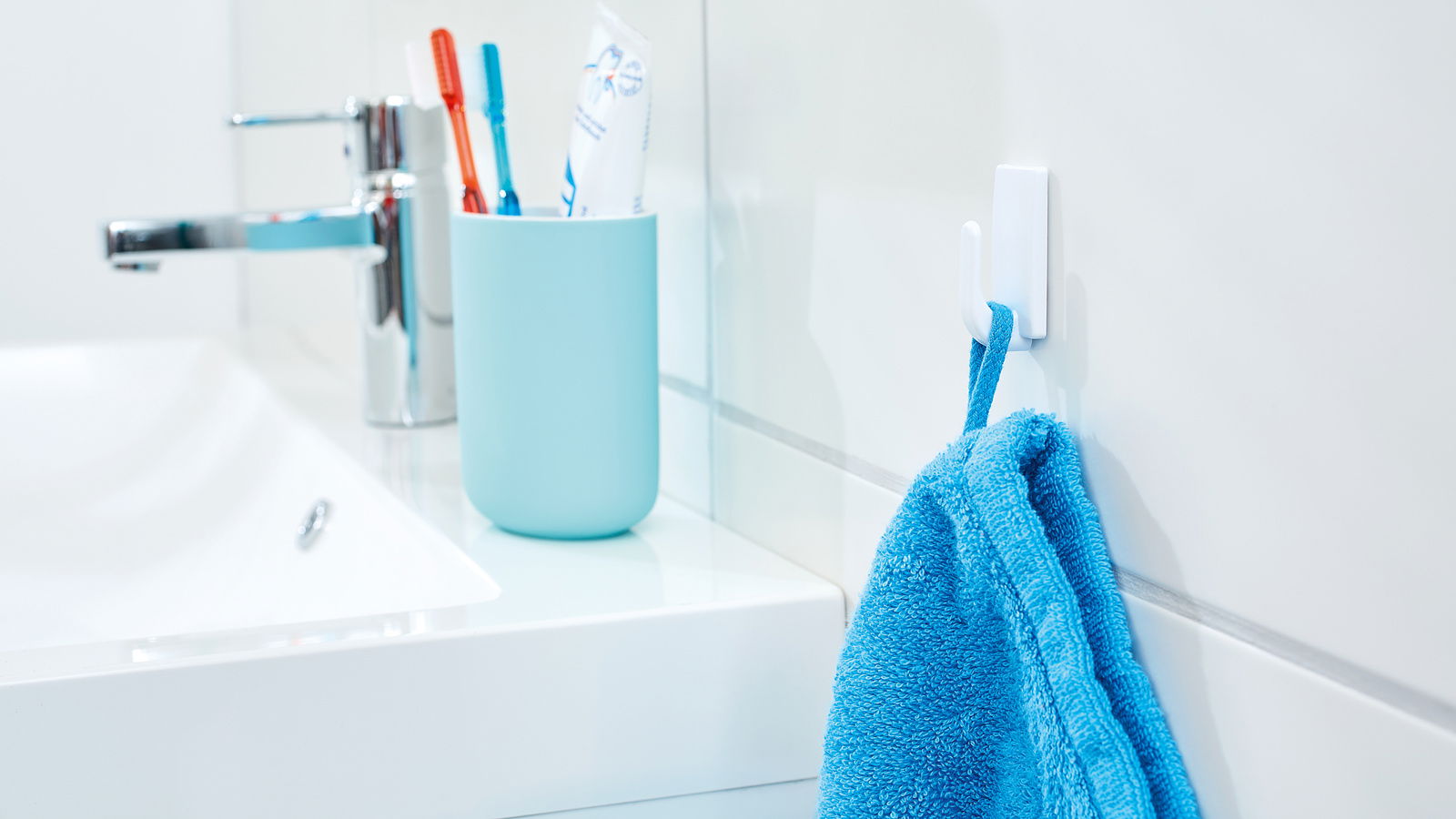 Tesa Powerstrips Haken 3-tlg. Badezimmeransicht mit einem weißen tesa Powerstrips Haken an der Wand, an dem ein blaues Handtuch hängt. Im Hintergrund sind ein Waschbecken, ein Wasserhahn und ein Becher mit Zahnbürsten zu sehen.