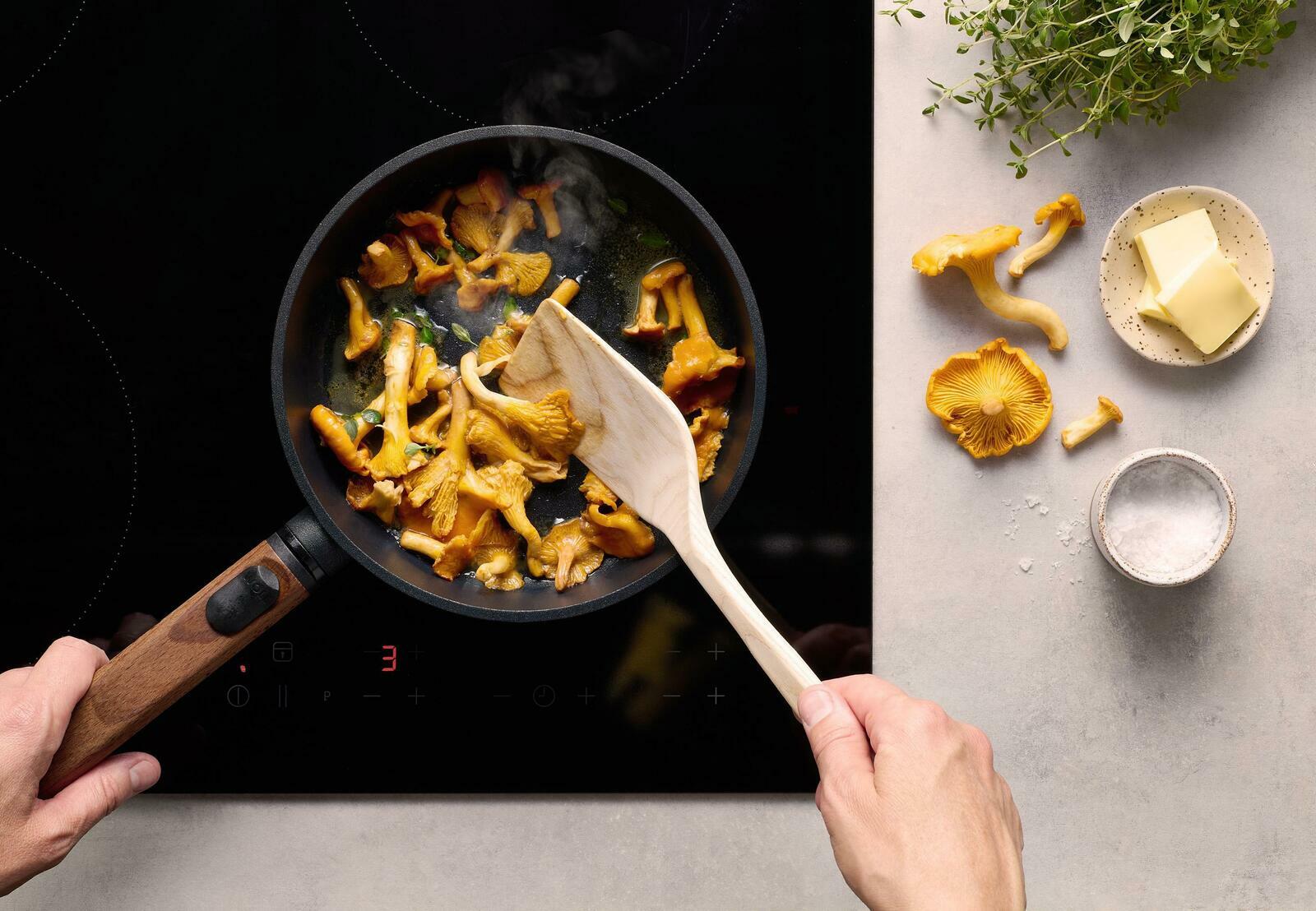 Obenansicht einer Guss-Flachpfanne mit Holzgriff, in der Pfifferlinge auf einem Herd gebraten werden. Daneben liegen Butter, Salz und frische Kräuter.
