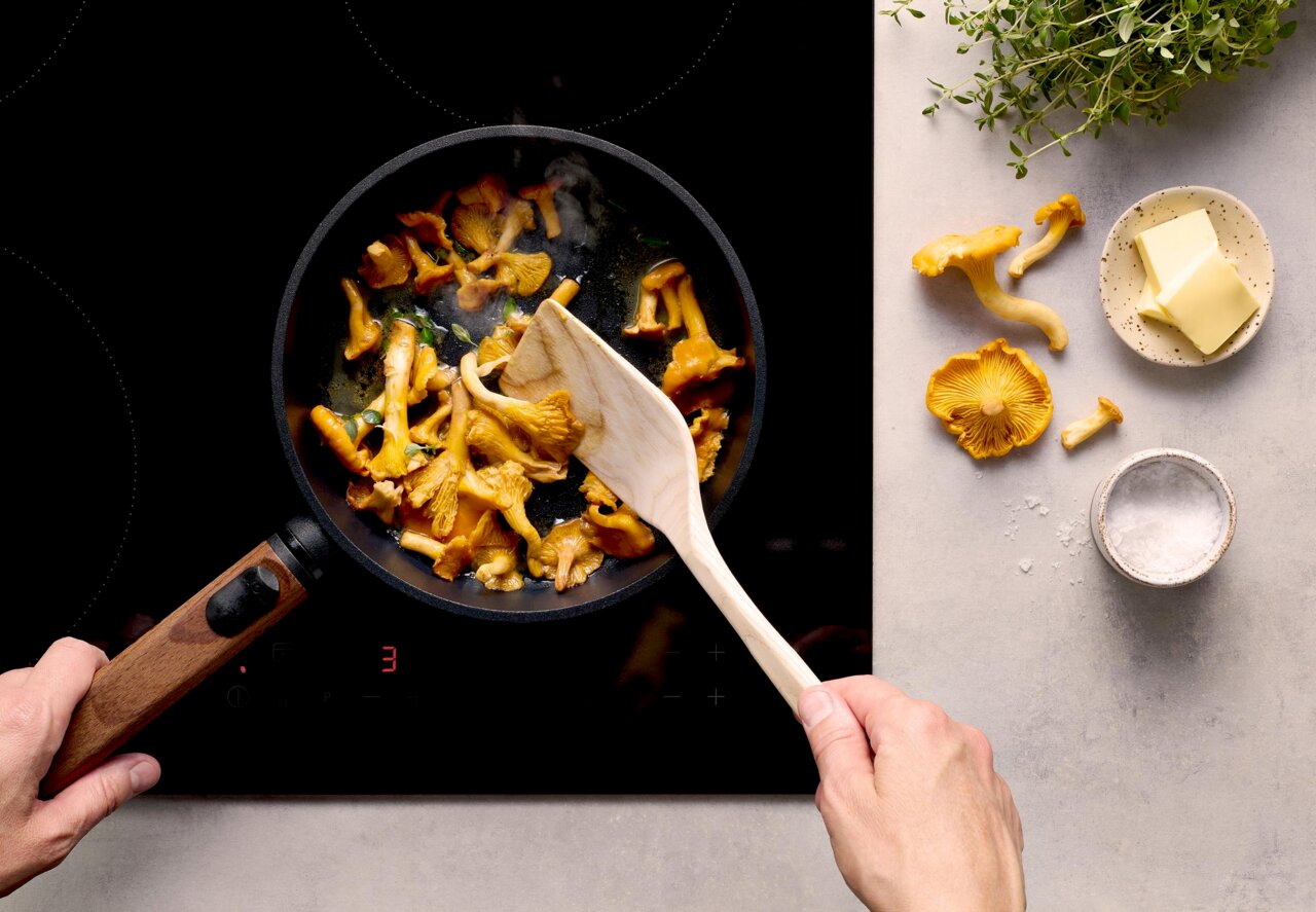 Woll Guss-Flachpfanne ECO LOGIC QXR Obenansicht einer Guss-Flachpfanne mit Holzgriff, in der Pfifferlinge auf einem Herd gebraten werden. Daneben liegen Butter, Salz und frische Kräuter.
