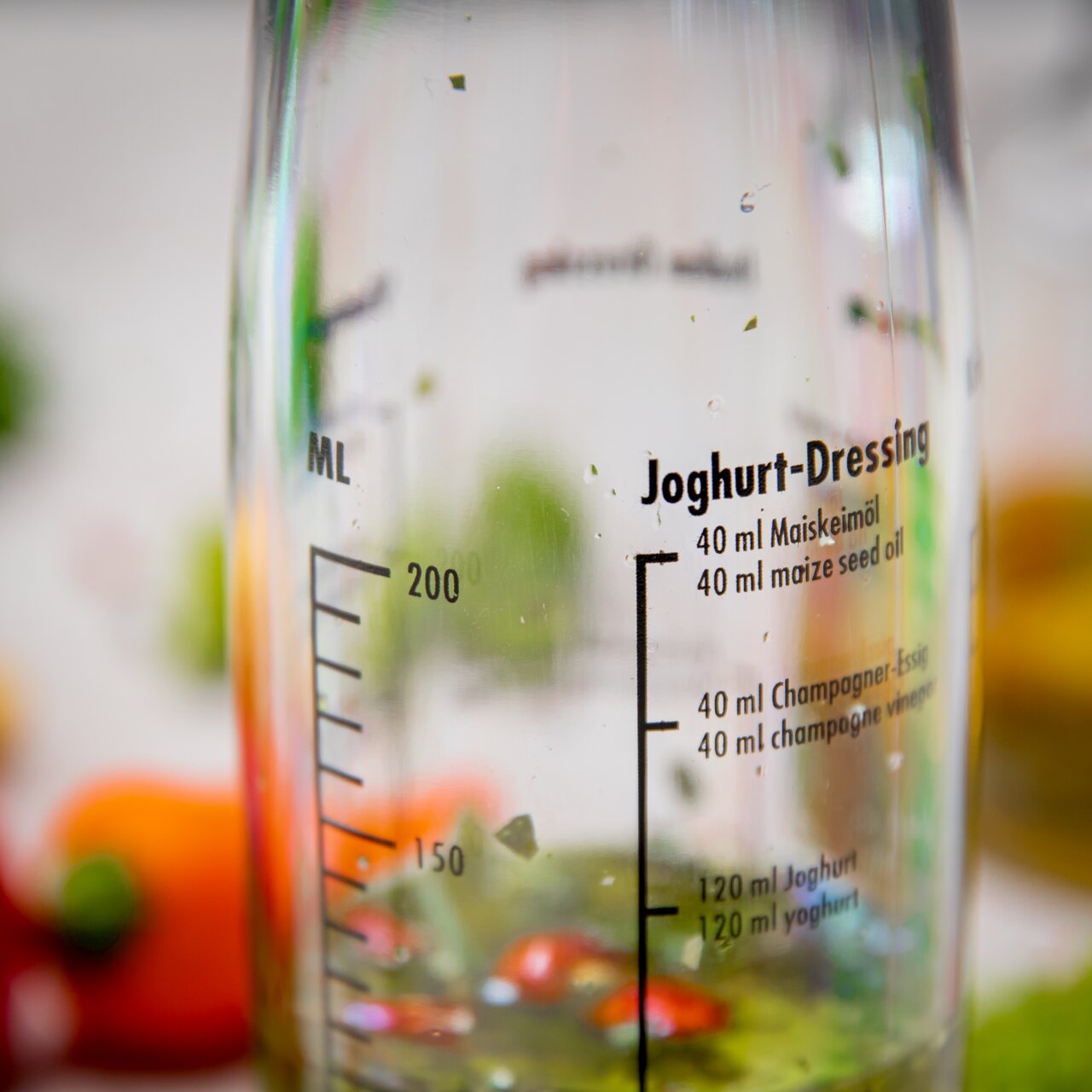 Nahaufnahme eines Dressing-Shakers mit Messskala und Rezept für Joghurt-Dressing auf der Glasoberfläche.